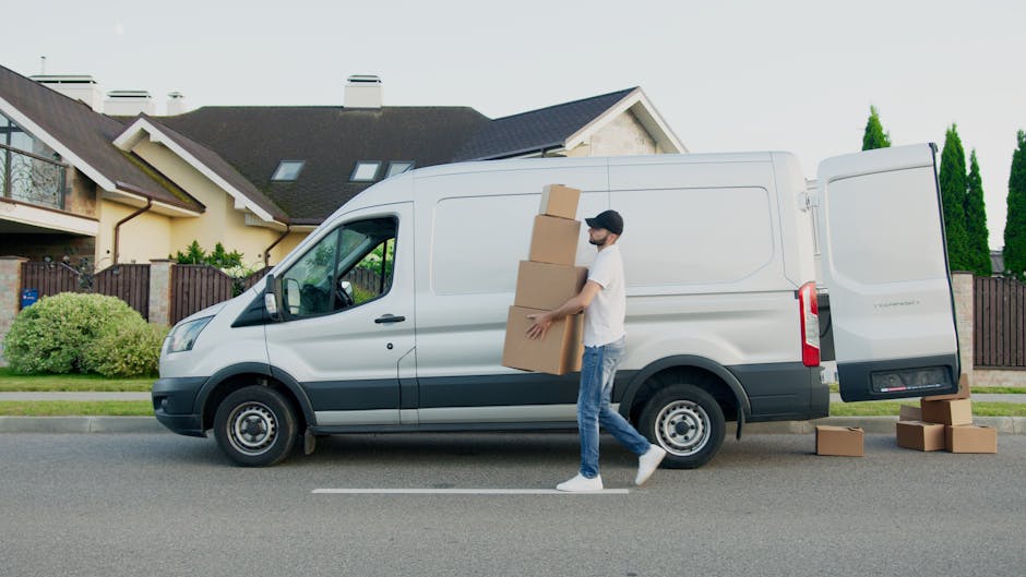 A man wearing a white T-shirt, light blue jeans, and a black cap is carrying three large cardboard boxes stacked one on top of the other while walking beside a white delivery van parked on a quiet residential street. The van has its rear doors open, with additional cardboard boxes on the ground nearby, indicating a home relocation or furniture transport process. The background features modern houses with sloped roofs, windows, and a garden with neatly trimmed bushes and tall, narrow trees, under bright natural light. Man and Van Kennington's vehicle is positioned close to the curb, suggesting an active loading or unloading scene typical of house removals or moving services. The environment appears calm and organized, appropriate for professional packing, moving, and transportation activities.