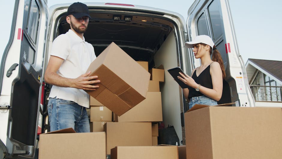 A young man with a beard, wearing a red backward cap, brown and beige jacket, checked shirt, and striped gloves, is sitting on the open tailgate of a black van. Inside the van, there are several cardboard moving boxes of various sizes, some stacked and others placed on the floor, all packed for a home relocation. The background shows the vehicle's interior, which is dark with visible padding and seat upholstery. The scene is outdoors, with natural lighting illuminating the man and the boxes, indicating an active loading process typical of furniture transport or packing and moving services, facilitated by Man and Van Kennington as part of their removals operations.