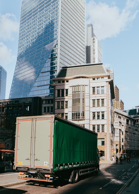 A large green-moving trailer attached to a commercial vehicle parked on a city street in front of modern high-rise office buildings and older architectural structures. The trailer is positioned on the pavement, with its rear doors securely closed, ready for furniture transport or packing materials during home relocation. The urban environment features clear blue skies and a few pedestrians, emphasizing the busy setting typical of professional removals and moving services like those provided by Man and Van Kennington. The scene captures the loading process, with the vehicle prepared for moving household belongings, emphasizing the importance of efficient logistics during furniture transport and packing and moving operations in a cityscape.