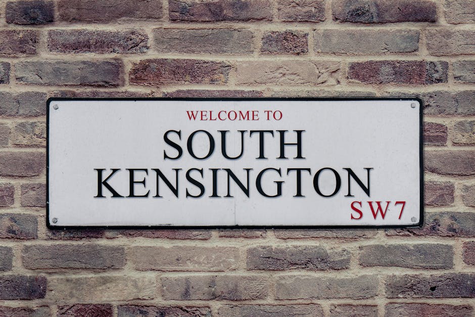 A rectangular white metal street sign with black and red text, mounted on a brick wall made of reddish-brown bricks with visible mortar joints. The sign reads 'Welcome to South Kensington' in large black letters, with 'SW7' in smaller red font at the bottom right corner, and the words 'Welcome to' in smaller red font above. The sign is positioned at chest height, and the surrounding brickwork provides a textured, urban backdrop typical of a residential or commercial property in South Kensington, London. The image captures the sign and the detailed brick wall for clarity and contextual realism relevant to house and property identification within a moving or relocation services setting.