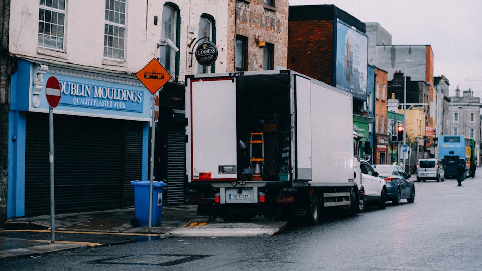 A large green-moving trailer attached to a commercial vehicle parked on a city street in front of modern high-rise office buildings and older architectural structures. The trailer is positioned on the pavement, with its rear doors securely closed, ready for furniture transport or packing materials during home relocation. The urban environment features clear blue skies and a few pedestrians, emphasizing the busy setting typical of professional removals and moving services like those provided by Man and Van Kennington. The scene captures the loading process, with the vehicle prepared for moving household belongings, emphasizing the importance of efficient logistics during furniture transport and packing and moving operations in a cityscape.