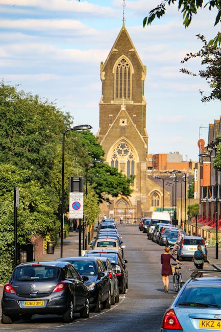A wide street scene showing a line of parked cars along the pavement with a woman walking her bicycle and another pedestrian nearby. The street is lined with black lampposts and trees, with residential buildings on the right side. In the background, a large historic stone church with Gothic-style arched windows and a tall, pointed steeple dominates the scene. The sky is mostly clear with some light clouds. The photo captures a typical urban environment during daylight hours, relevant to home relocation or moving services such as those offered by Man and Van Kennington, illustrating an area where furniture transport and loading processes could take place during a house removal.