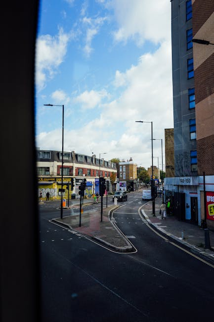 View of a busy urban street scene seen through a partially open window, featuring a curved road with a white van and other vehicles, tall modern buildings on the right, and a row of multi-story shops across the street with signage, some with yellow and black advertising banners. The sidewalk nearby hosts a few pedestrians, and street lamps line both sides of the road. The sky overhead is partly cloudy with patches of blue. This scene illustrates the typical environment encountered during home relocation or furniture transport near Kennington and Vauxhall, where Man and Van Kennington provides removals services, including loading and unloading processes within urban streets.