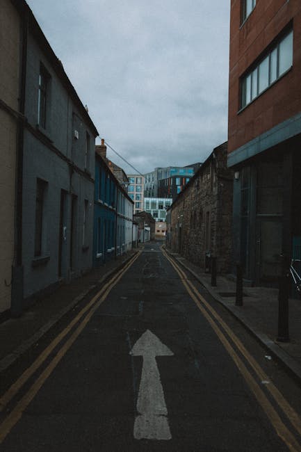 A narrow urban street with a single white arrow painted on the asphalt pointing forward, flanked by two rows of buildings with contrasting facades—on the left, older, weathered structures with small windows, and on the right, modern, brick and glass office or apartment buildings. The street is lined with black bollards on the sidewalk edges, and the sky above is overcast, creating dim lighting conditions. In the distance, a moving van or large vehicle may be visible, indicating a home relocation or furniture transport process. This scene captures the typical environment where Man and Van Kennington provides house removals, highlighting the tight street context and the need for careful, professional loading and unloading of furniture, boxes, and packing materials involved in residential moves.