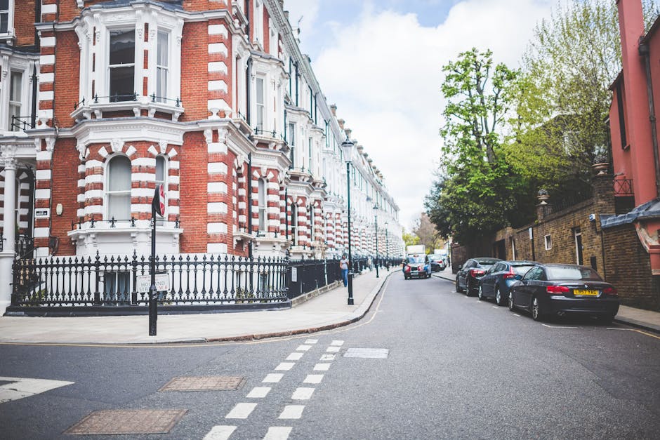 A street scene on Kennington Park Road showing a row of historic red-brick and white Victorian terrace houses with detailed architectural features, including bay windows and decorative mouldings. The foreground features a paved road with white dashed and solid lines, and a small pedestrian crossing. To the left, a Victorian-style cast iron fence surrounds a property with a sign indicating parking restrictions. Several parked cars, including a black sedan and a silver hatchback, are aligned along the curb on the right side. On the sidewalk, [COMPANY_NAME] is engaged in a house removal process, with visible cardboard boxes, wrapped furniture, and packing materials near the entrance of one of the houses, ready for transport. A van is parked on the street, with a portion of its loading area visible. The scene is illuminated by natural daylight, with a clear sky and some green trees providing additional context for a typical residential area in London focused on home relocation and furniture transport activities.