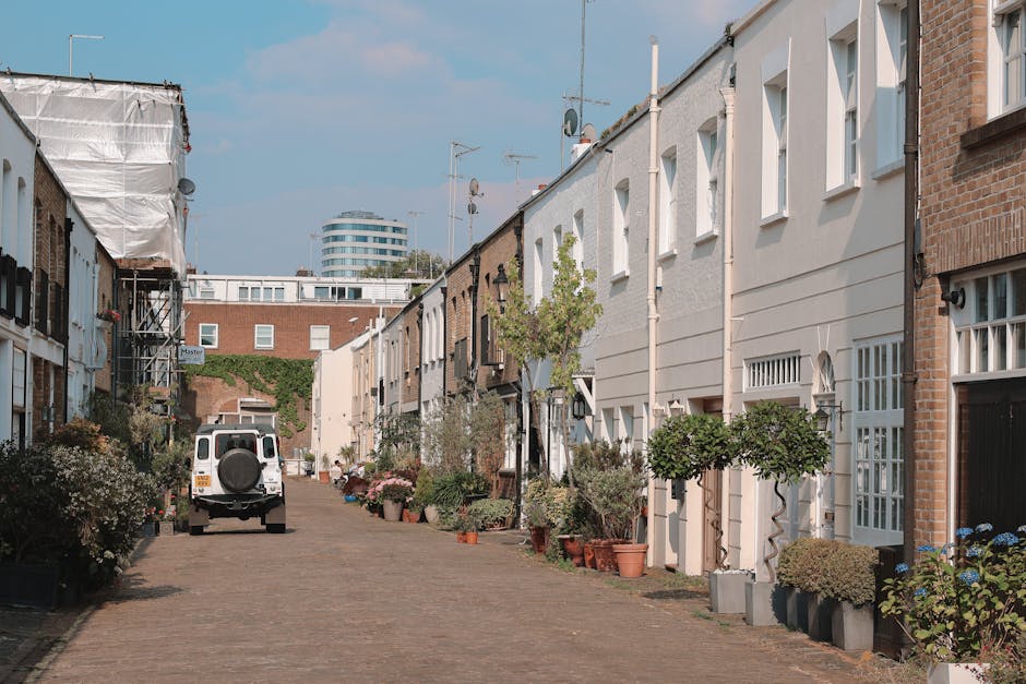 A narrow urban street with a single white arrow painted on the asphalt pointing forward, flanked by two rows of buildings with contrasting facades—on the left, older, weathered structures with small windows, and on the right, modern, brick and glass office or apartment buildings. The street is lined with black bollards on the sidewalk edges, and the sky above is overcast, creating dim lighting conditions. In the distance, a moving van or large vehicle may be visible, indicating a home relocation or furniture transport process. This scene captures the typical environment where Man and Van Kennington provides house removals, highlighting the tight street context and the need for careful, professional loading and unloading of furniture, boxes, and packing materials involved in residential moves.