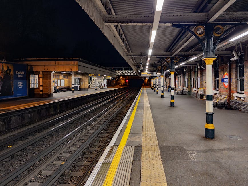 A view of a London Underground railway station platform at night, showing a row of brick and metal safety posts painted in yellow and black, supported by decorative cast iron columns with yellow accents, and a canopy overhead. The platform is illuminated by overhead lighting, with visible tactile paving near the edge of the platform for safety. To the left, there are benches, posters, and a person seated against the wall. The railway tracks run parallel to the platform, extending into the distance, with a station building visible on the opposite side. This setting depicts a typical scene during home relocation or furniture transport, where items are prepared for loading onto a vehicle, supported by professional removals services like Man and Van Kennington, and the platform environment is suitable for efficient loading and packing of boxes and furniture for house moves.