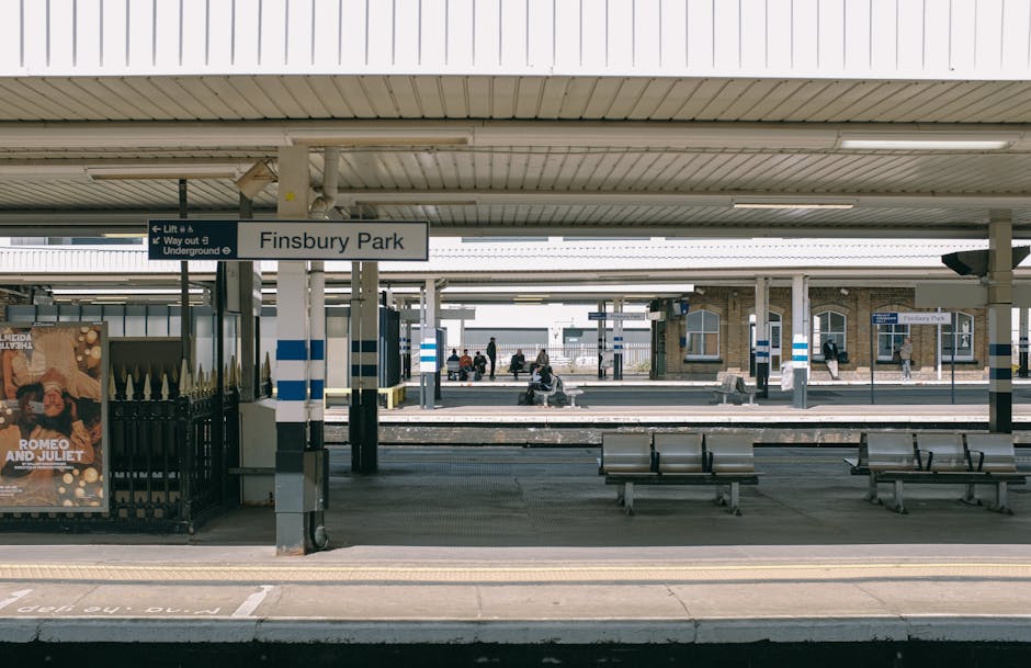 Finsbury Park railway station platform showing multiple seating benches and structural columns supporting the overhead canopy. A sign displaying 'Finsbury Park' is mounted on a column in the center of the image. Several passengers are visible seated on the benches and waiting, some reading or using electronic devices, while others stand near the platform edge. The platform surface is concrete with tactile paving strips along the edge for safety. A poster advertising the play 'Romeo and Juliet' is attached to a fence on the left side. The station environment includes a mixture of natural daylight and shadow from the canopy, with a background showing station buildings and additional train tracks. This setting reflects a typical urban railway station environment, relevant to home relocation or furniture transport logistics with Man and Van Kennington occasionally handling moving and packing services for clients near the Kennington area.