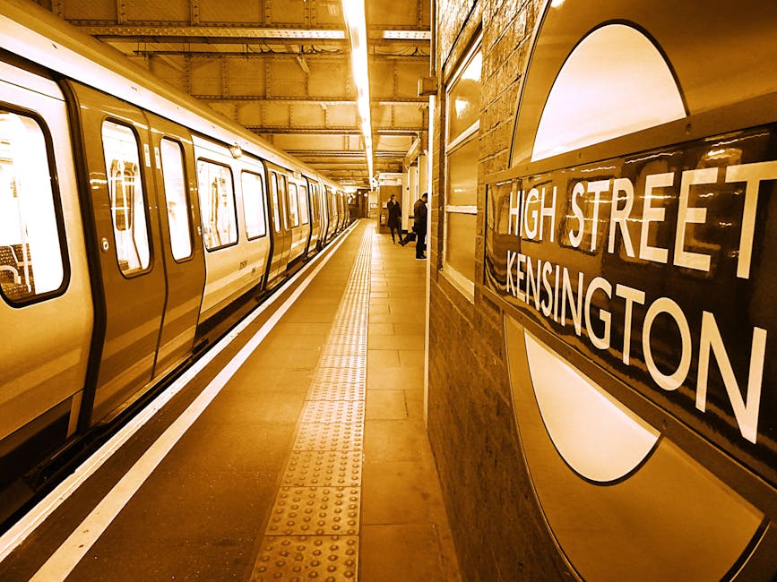 A view of a London Underground railway station platform at night, showing a row of brick and metal safety posts painted in yellow and black, supported by decorative cast iron columns with yellow accents, and a canopy overhead. The platform is illuminated by overhead lighting, with visible tactile paving near the edge of the platform for safety. To the left, there are benches, posters, and a person seated against the wall. The railway tracks run parallel to the platform, extending into the distance, with a station building visible on the opposite side. This setting depicts a typical scene during home relocation or furniture transport, where items are prepared for loading onto a vehicle, supported by professional removals services like Man and Van Kennington, and the platform environment is suitable for efficient loading and packing of boxes and furniture for house moves.