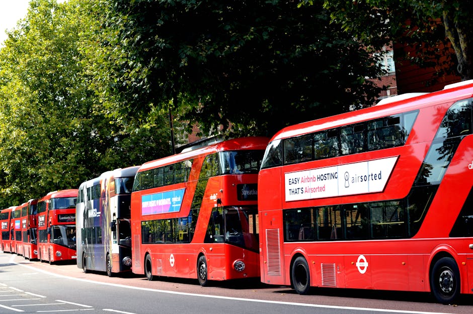 A line of red double-decker buses parked on a street in Lambeth, with some buses displaying advertisements on their sides. The scene is set outdoors during daytime, with a large leafy green tree partially shaded the buses and a building with brick and glass facades visible in the background. The buses are aligned along the curb, with the first bus in the foreground clearly showing the London transport roundel symbol and tinted windows, while the others extend into the distance. The pavement is visible, and there are no pedestrians in view. This image captures a typical scene of public transportation in Lambeth, relevant to home relocation or furniture transport services such as those offered by Man and Van Kennington, especially during moving logistics involving busy urban environments.