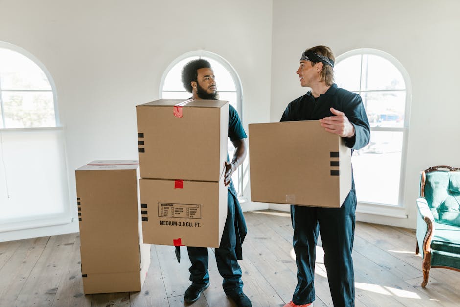 Two movers from Man and Van Kennington are inside a well-lit room with light-colored walls and wooden flooring, preparing for home relocation by handling moving boxes. The person on the left, with an afro hairstyle and casual clothing, is holding a medium-sized cardboard box that has black printed markings indicating its size, along with red packing tape. The individual on the right, wearing a black tracksuit and a headband, is carrying a similarly sized cardboard box with tape, gesturing as if coordinating the loading process. Behind them, three additional cardboard boxes are positioned on the floor, with one box partially obscured by the mover on the left, suggesting they are in the midst of packing or loading items for a furniture transport or moving service. Large arched windows in the background allow natural light to illuminate the scene, emphasizing the indoor setting, where careful handling of packing materials is part of the moving process managed by professional services like Man and Van Kennington, ensuring efficiency in furniture transport and packing during a house move.