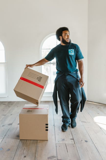 A man with an Afro hairstyle and beard, dressed in a navy-blue t-shirt and dark trousers, is inside a well-lit room with white walls and wooden flooring. He is holding a medium-sized cardboard box wrapped with red packing tape in his left hand, preparing to place or lift it onto another slightly larger cardboard box positioned on the floor. Behind him, a large arched window allows natural light into the space, revealing an outdoor scene. The scene depicts a professional home relocation or furniture transport process, with the man engaged in packing and moving activities associated with house removals, supported by the presence of multiple cardboard boxes and the indoor environment typical of a property undergoing a move. Man and Van Kennington, offering removals services, is referenced through the context.