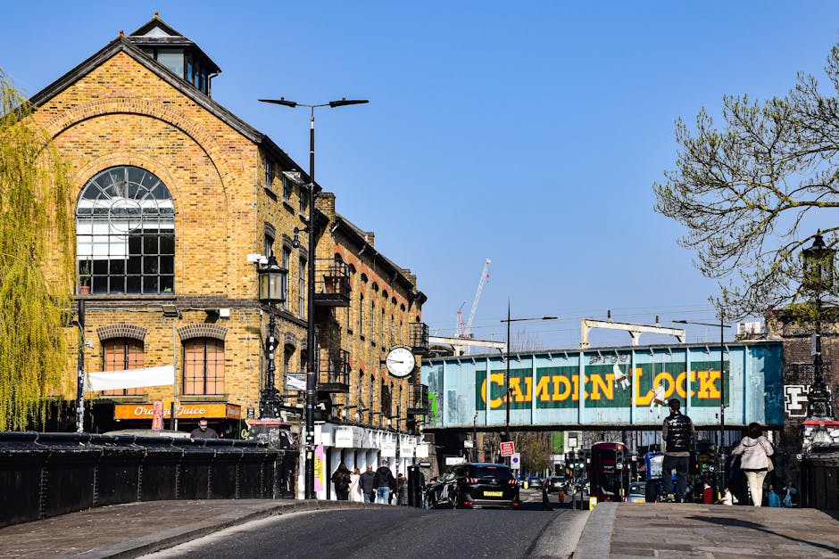 A street scene in Kennington, London, near The Oval, featuring a row of brick buildings with large arched windows and a clock mounted on a black pole. In the foreground, pedestrians are walking along the sidewalk, some carrying bags, with vehicles parked along the roadside. A blue railway bridge with the words 'CAMDEN LOCK' painted in large yellow letters spans the street, with an industrial aesthetic. Nearby, a crane is visible in the distance against a clear blue sky, indicating ongoing construction. The street is illuminated by black street lamps, and a tall lamppost with dual overhead lights is centered above the scene. On the left, part of a tree with budding leaves hangs over the sidewalk, providing a touch of greenery. The environment appears busy with active foot and vehicle traffic, capturing a typical day in an urban area involved in home relocation or moving logistics where the Man and Van Kennington team might operate, including furniture transport and packing and moving processes within this lively street setting.