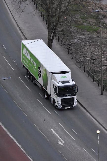 An overhead view of a white delivery truck parked on a city street, with the logo and branding for ASDA Money displayed on the side in green and black. The truck is positioned along a paved road with clear lane markings, next to a sidewalk separated by a low fence and a row of leafless trees. The surrounding environment includes a patch of bare ground or park area and some distant urban infrastructure. This scene exemplifies logistics and vehicle aspects of home relocation services, such as furniture transport and packing and moving operations managed by companies like Man and Van Kennington, supporting efficient house removals near SE11 Kennington and The Oval areas.