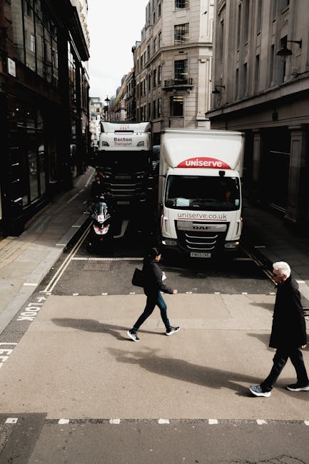 A street scene showing a narrow urban road with tall, multi-storey buildings on both sides. Two white moving vans with the branding 'uniserve' are parked perpendicular to the curb, partly blocking the road, indicating active loading or unloading during a house relocation. A black scooter is parked beside the vans, close to the pavement. In the foreground, a man and a woman are walking across a pedestrian crossing; the woman is carrying a backpack and wearing casual clothing, while the older man is dressed in a dark suit and has white hair. The scene is illuminated by natural daylight, with overcast skies visible in the background. The environment suggests a busy city centre location, with the presence of moving equipment and vehicles relevant to the process of packing and transporting furniture and household items for a home relocation. The image highlights the logistical aspects of short-notice removals services, such as those provided by Man and Van Kennington, in an urban setting.